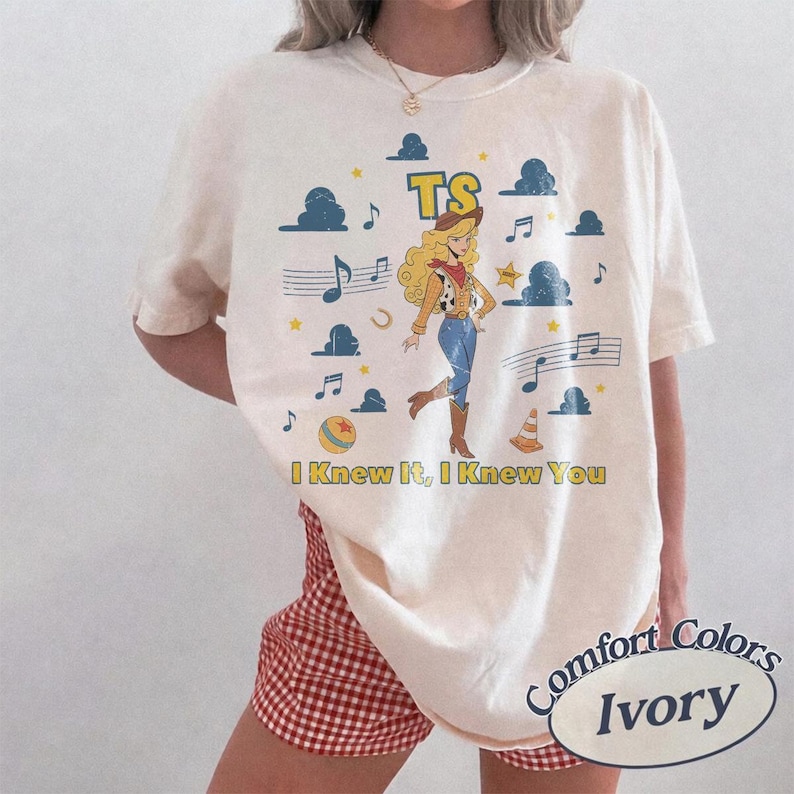 May include: An ivory short-sleeve t-shirt featuring a graphic of a blonde cowgirl character, musical notes, stars, clouds, and a ball. The text reads TS and I Knew It, I Knew You. The shirt has a crew neckline and a relaxed fit, paired with red gingham shorts.