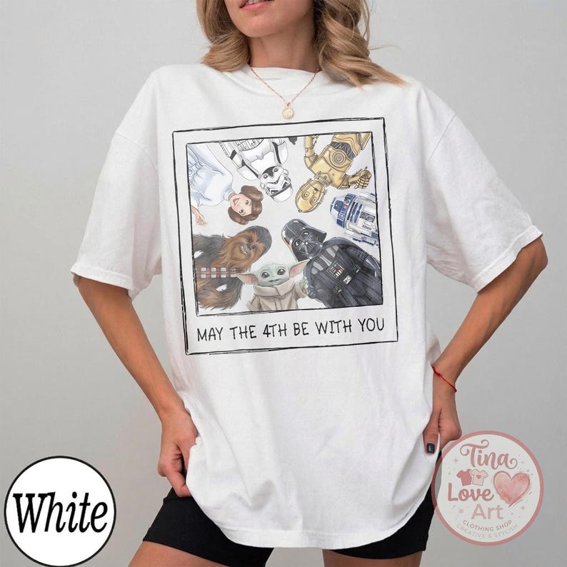 May include: White t-shirt featuring a framed illustration of Star Wars characters, including Darth Vader and Baby Yoda. The text "MAY THE 4TH BE WITH YOU" is printed below the image. The shirt has short sleeves.