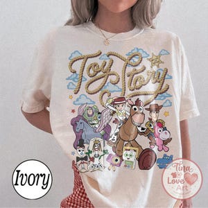 Op de afbeelding: Een ivoorkleurig t-shirt met een grafische print van Toy Story-personages, waaronder Buzz Lightyear, Jessie, Woody, Bullseye en anderen. De tekst Toy Story staat in een touwachtig lettertype boven de personages, met blauwe wolken en sterren.