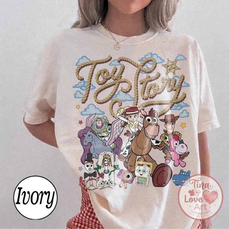 Op de afbeelding: Een ivoorkleurig t-shirt met een grafische print van Toy Story-personages, waaronder Buzz Lightyear, Jessie, Woody, Bullseye en anderen. De tekst Toy Story staat in een touwachtig lettertype boven de personages, met blauwe wolken en sterren.