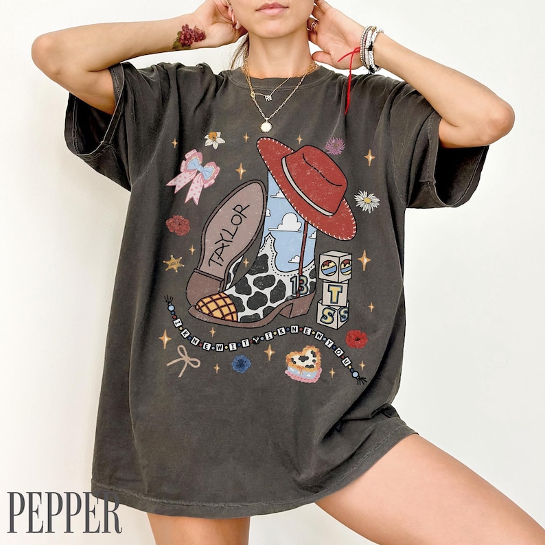 May include: A charcoal grey t-shirt featuring a graphic print of a cow-print cowboy boot, a red hat, a bow, and blocks with the letters T, S, and the number 13. The design includes the text TAYLOR and I KNEW YOU WERE TROUBLE on a beaded string.