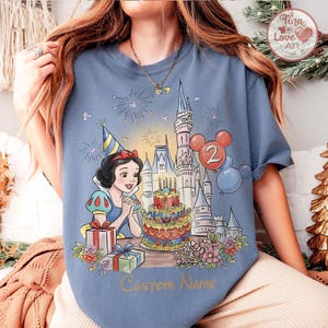May include: A pale blue t-shirt with a Snow White graphic, a birthday cake, a castle, and balloons. The design includes the number 2, gift boxes, and the text "Custom Name". The shirt has a classic crew neck.