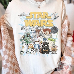 May include: Cream-colored t-shirt featuring a graphic of Star Wars characters and the Star Wars logo in yellow. The design includes characters like Darth Vader, Yoda, and Boba Fett, with a light blue background.