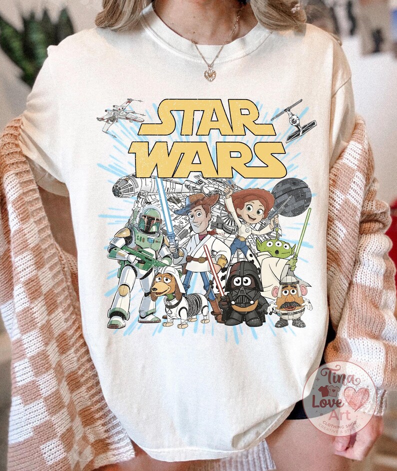 May include: Cream-colored t-shirt featuring a graphic of Star Wars characters and the Star Wars logo in yellow. The design includes characters like Darth Vader, Yoda, and Boba Fett, with a light blue background.