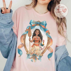 May include: Pink t-shirt featuring a framed illustration of a Polynesian woman holding a spear, with a pig and rooster. The frame is adorned with blue and orange ribbons. The shirt has the text "Tina Love Art".