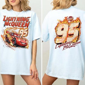 May include: A light blue t-shirt featuring a graphic of the red race car Lightning McQueen with the text Lightning McQueen and the number 95 on the front, and a large number 95 surrounded by flames with the text Lightning McQueen on the back.