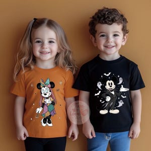Op de afbeelding: Twee kinderen dragen Halloween-t-shirts. Het ene kind draagt een oranje shirt met een Minnie Mouse-ontwerp in een patchworkjurk. Het andere kind draagt een zwart shirt met een Mickey Mouse-ontwerp in een Jack Skellington-kostuum, omringd door vleermuizen en geesten.