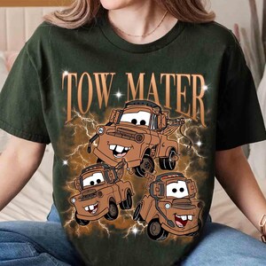 Może przedstawiać: Ciemnozielony t-shirt z kr&oacute;tkim rękawem z nadrukiem graficznym przedstawiającym trzy brązowe holowniki. Napis TOW MATER jest wydrukowany dużą beżową czcionką nad pojazdami, w otoczeniu brązowego wzoru błyskawic z małymi białymi gwiazdkami.