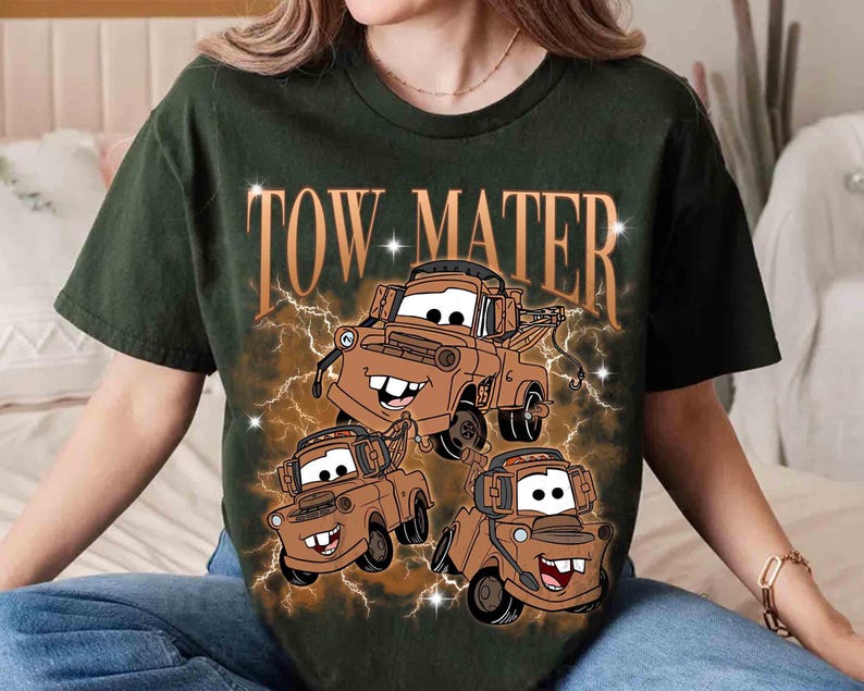 Może przedstawiać: Ciemnozielony t-shirt z kr&oacute;tkim rękawem z nadrukiem graficznym przedstawiającym trzy brązowe holowniki. Napis TOW MATER jest wydrukowany dużą beżową czcionką nad pojazdami, w otoczeniu brązowego wzoru błyskawic z małymi białymi gwiazdkami.