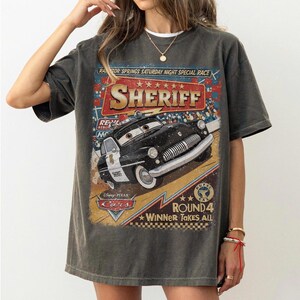 May include: A charcoal grey t-shirt featuring a graphic print of the character Sheriff from the Disney Pixar Cars movie. The design includes the text Radiator Springs Saturday Night Special Race, Sheriff, Disney Pixar Cars, and Round 4 Winner Takes All.