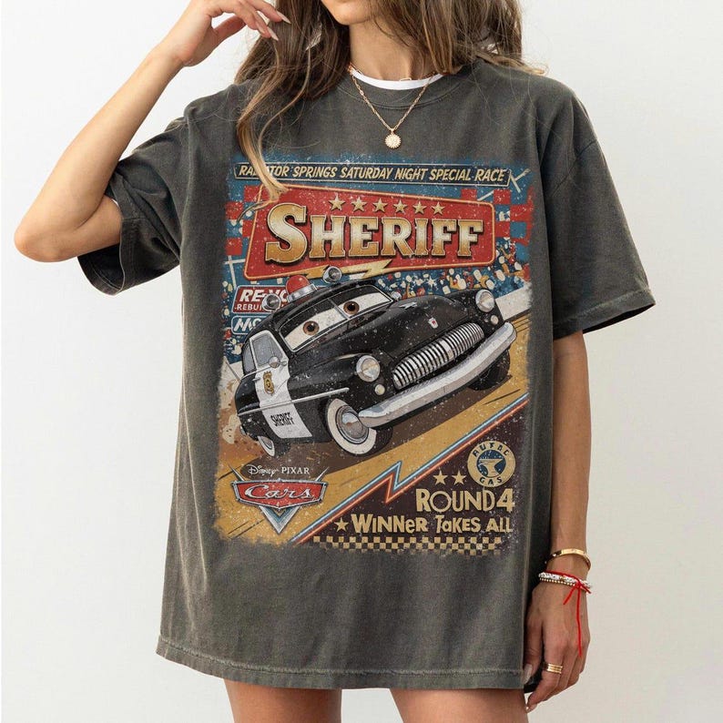 May include: A charcoal grey t-shirt featuring a graphic print of the character Sheriff from the Disney Pixar Cars movie. The design includes the text Radiator Springs Saturday Night Special Race, Sheriff, Disney Pixar Cars, and Round 4 Winner Takes All.