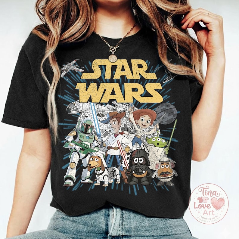 May include: Black t-shirt featuring a graphic of Star Wars characters, including Darth Vader, Woody, and Buzz Lightyear. The words "STAR WARS" are in yellow block letters above the characters. A Tina Love Art logo is in the bottom right corner.
