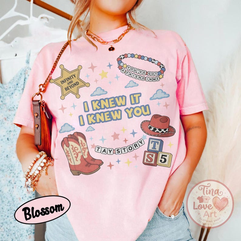May include: A light pink short-sleeve t-shirt featuring a graphic print with the text I KNEW IT I KNEW YOU, TAY STORY, INFINITY AND BEYOND, and YOU'VE GOT A FRIEND IN ME. The design includes a sheriff badge, cowboy boots, a hat, and blocks.