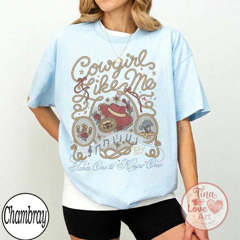 Pu&ograve; includere: Una t-shirt azzurro chiaro con una stampa grafica bordata da corde, un cappello da cowboy rosso, un ferro di cavallo e note musicali. Il testo recita "Cowgirl like Me" e "Takes One to Know One" in corsivo, circondato da stelline e motivi floreali.