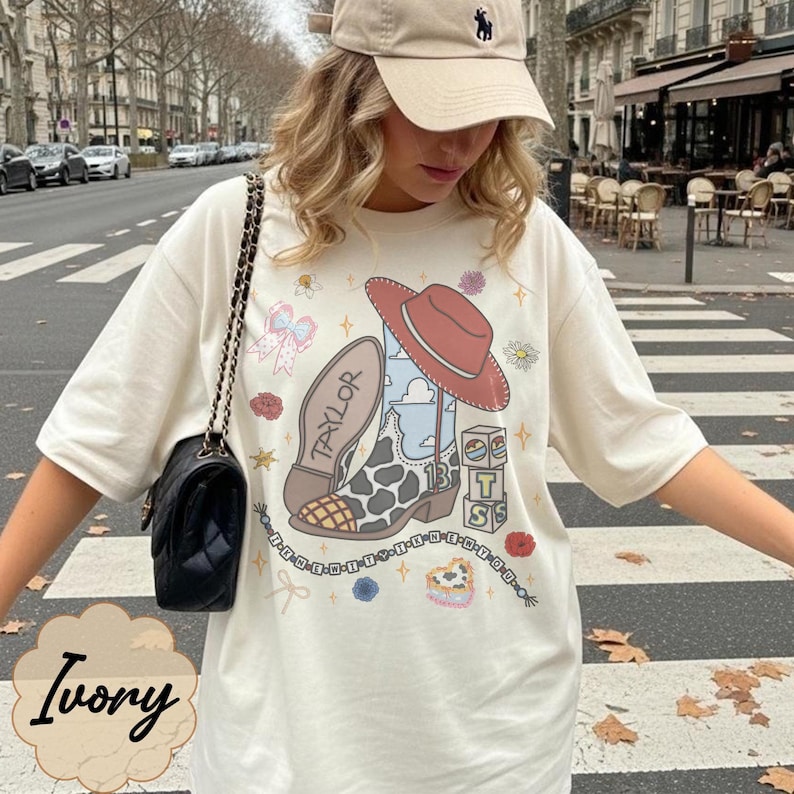 May include: An ivory t-shirt featuring a graphic print of a cowboy boot with a cow print pattern, a red hat, and the name Taylor. The design includes blocks with the letters T, S, and the number 13, surrounded by small floral and star icons.