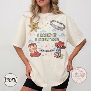 May include: An ivory short-sleeve t-shirt featuring a graphic print with the text I KNEW IT I KNEW YOU, TAY STORY, and INFINITY AND BEYOND. The design includes illustrations of a sheriff badge, a beaded bracelet, cowboy boots, a hat, and blocks.
