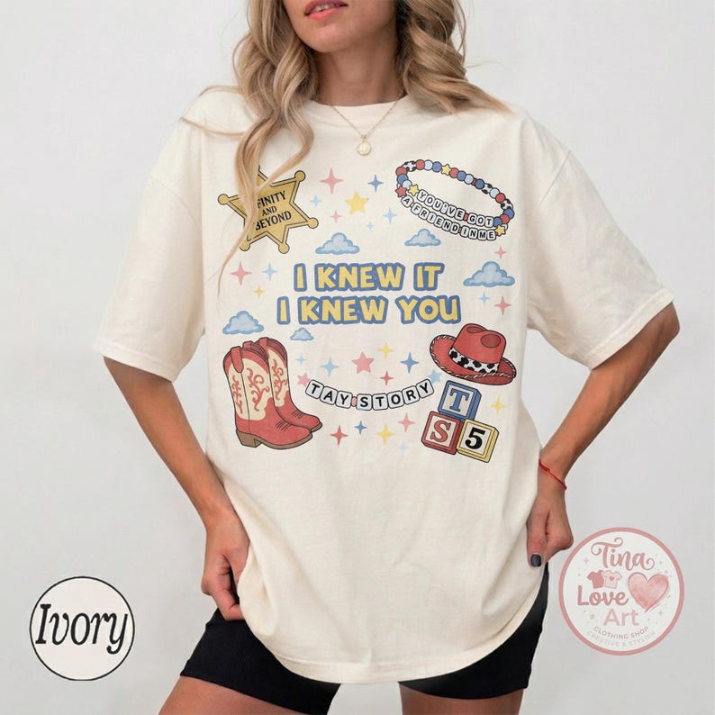 May include: An ivory short-sleeve t-shirt featuring a graphic print with the text I KNEW IT I KNEW YOU, TAY STORY, and INFINITY AND BEYOND. The design includes illustrations of a sheriff badge, a beaded bracelet, cowboy boots, a hat, and blocks.