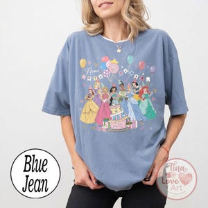 May include: Blue jean colored t-shirt featuring a birthday party design with Disney princesses, balloons, and a cake. The number 5 is displayed on the shirt, along with the word "Name". The shirt has a round logo that says "Blue Jean".