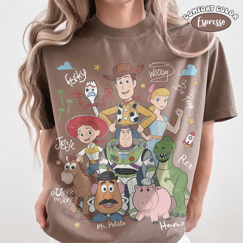 May include: An espresso-colored t-shirt featuring a graphic print of Toy Story characters including Woody, Buzz Lightyear, Jessie, Bullseye, Rex, Hamm, Mr. Potato Head, Bo Peep, and Forky, with handwritten-style character names and small star icons.