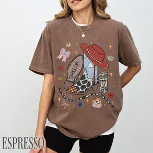 May include: A brown short-sleeve t-shirt featuring a graphic print of a cowboy boot with a cow print pattern, a red hat, a pink bow, and blocks with the letters T, S, and the number 13. The text TAYLOR is on the boot, and a beaded string reads THE NEW IT KNEW N YOU.