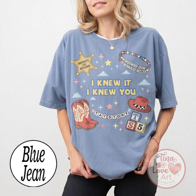 May include: A blue jean colored t-shirt featuring a graphic print with a gold sheriff star, a beaded bracelet, a red cowboy boot, a red cowboy hat, and blocks with the text T S 5. The shirt includes the text I KNEW IT I KNEW YOU.