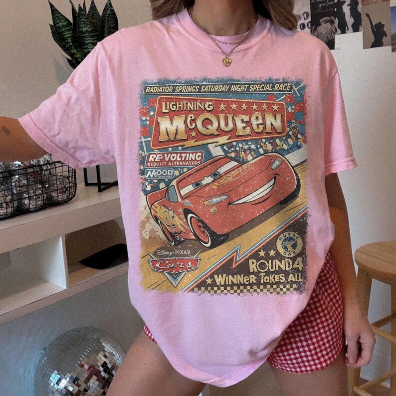 May include: A light pink short-sleeve t-shirt featuring a graphic print of the character Lightning McQueen from the movie Cars. The design includes the text Radiator Springs Saturday Night Special Race, Lightning McQueen, Re-Volting, and Round 4.