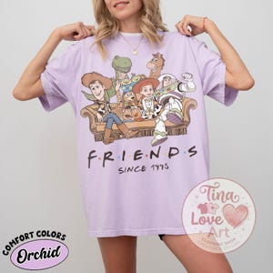 Peut inclure: Un t-shirt violet clair avec une impression graphique repr&eacute;sentant divers personnages de jouets anim&eacute;s assis sur un canap&eacute;. Sous les personnages, le texte "FRIENDS SINCE 1995" est &eacute;crit en noir. Le t-shirt a des manches courtes et un col rond.