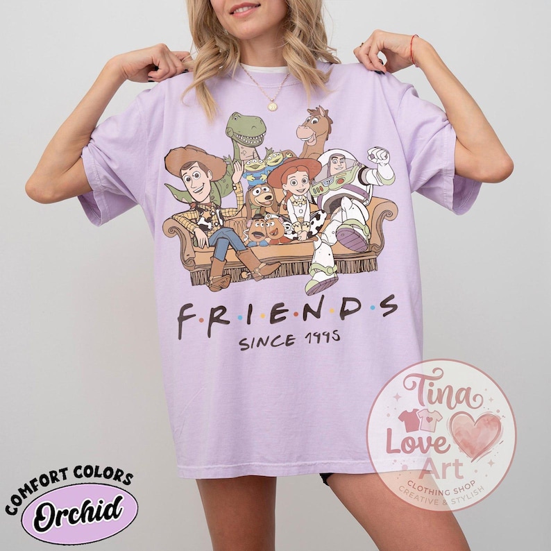 Peut inclure: Un t-shirt violet clair avec une impression graphique repr&eacute;sentant divers personnages de jouets anim&eacute;s assis sur un canap&eacute;. Sous les personnages, le texte "FRIENDS SINCE 1995" est &eacute;crit en noir. Le t-shirt a des manches courtes et un col rond.