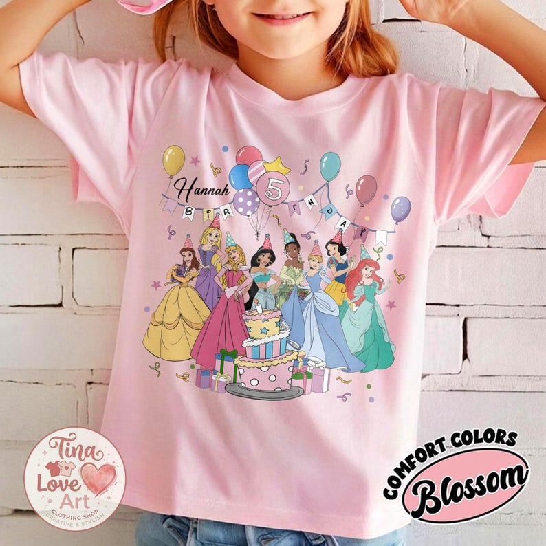 May include: Pink t-shirt featuring a colorful graphic of Disney princesses, balloons, and a birthday cake. The shirt has the name "Hannah" and the number "5" printed on it. The shirt is from the "Comfort Colors" brand.
