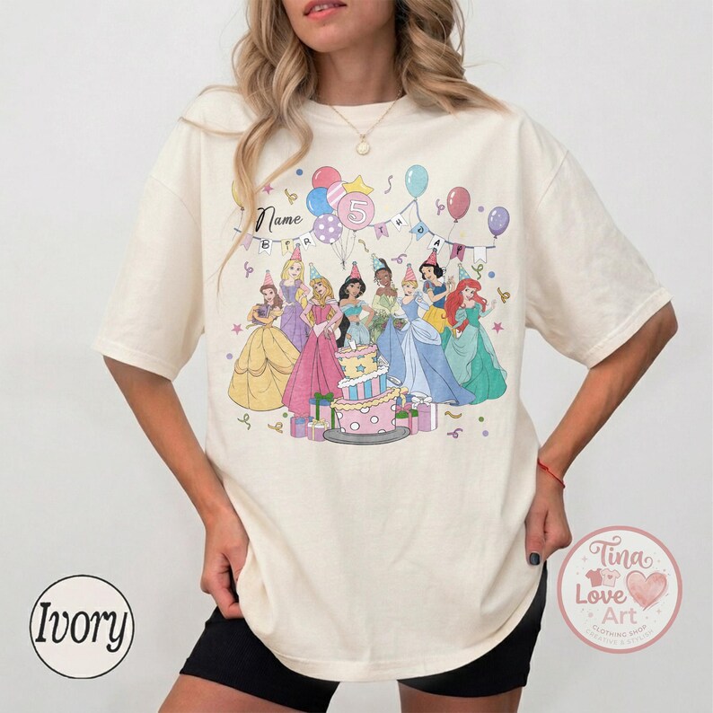 May include: Ivory-colored t-shirt featuring a colorful graphic of Disney princesses around a birthday cake. The design includes balloons, party hats, and the number 5. The word "Name" is also visible on the shirt.