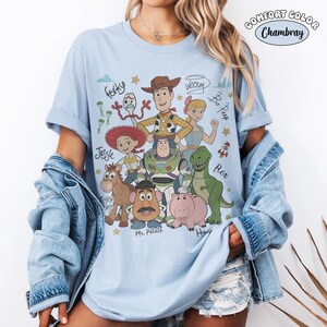 May include: A light blue t-shirt featuring a graphic print of Toy Story characters including Woody, Buzz Lightyear, Jessie, Rex, Hamm, Mr. Potato Head, Bo Peep, Forky, and Bullseye, with their names written in cursive script and small star icons.