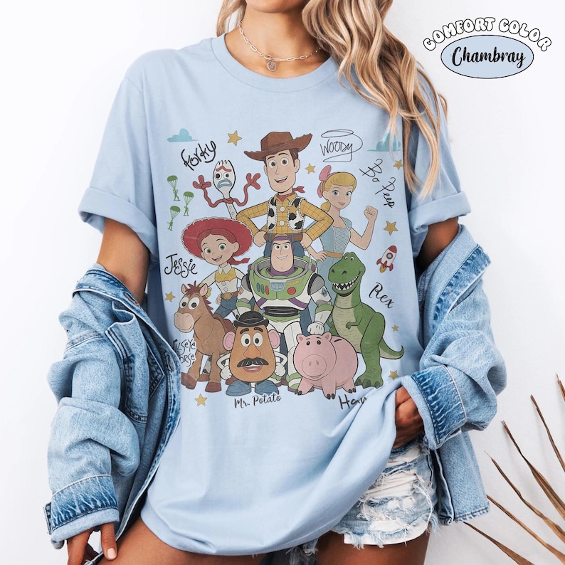 May include: A light blue t-shirt featuring a graphic print of Toy Story characters including Woody, Buzz Lightyear, Jessie, Rex, Hamm, Mr. Potato Head, Bo Peep, Forky, and Bullseye, with their names written in cursive script and small star icons.