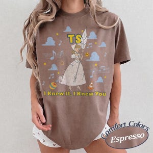 May include: A brown short-sleeve t-shirt featuring a graphic of a cartoon character in a white dress holding a large white feather. The design includes blue clouds, yellow stars, musical notes, a ball, a traffic cone, and the text TS I Knew It, I Knew You.