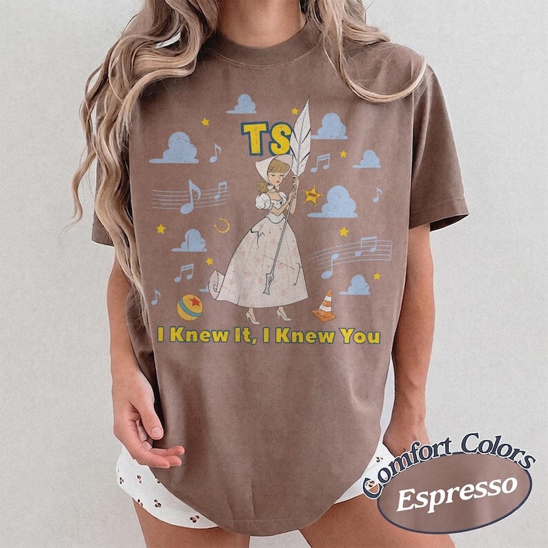 May include: A brown short-sleeve t-shirt featuring a graphic of a cartoon character in a white dress holding a large white feather. The design includes blue clouds, yellow stars, musical notes, a ball, a traffic cone, and the text TS I Knew It, I Knew You.