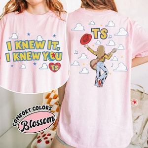 K&ouml;nnte beinhalten: Ein hellrosa T-Shirt mit einem Grafikdruck. Die Vorderseite zeigt den Text I KNEW IT, I KNEW YOU in Gelb mit blauen Umrandungen und ein rotes Herz mit TS. Die R&uuml;ckseite zeigt eine Person mit Gitarre, rotem Hut und einer Hose mit Kuhmuster.