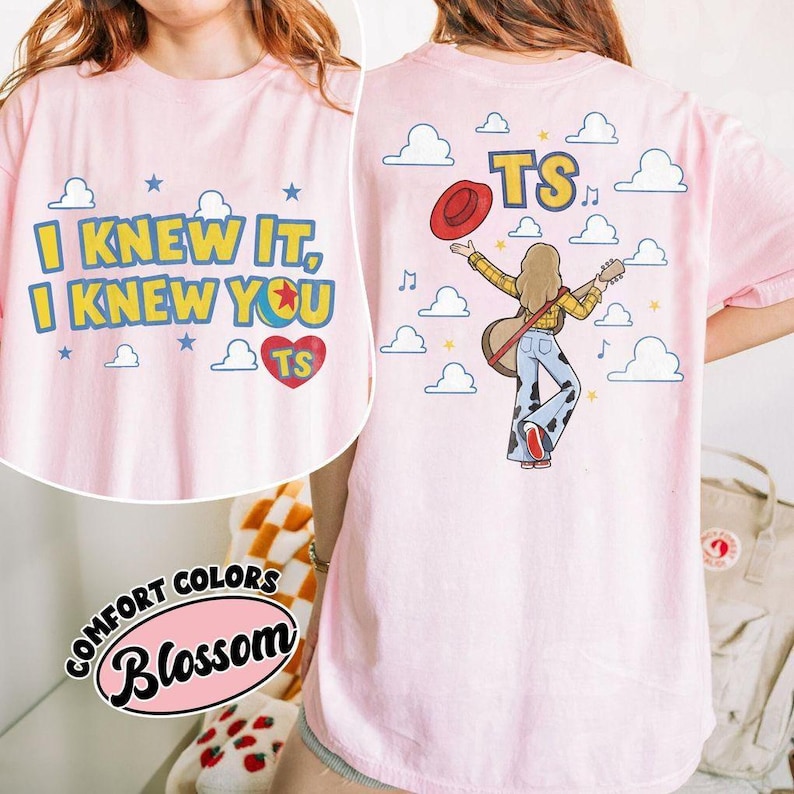 K&ouml;nnte beinhalten: Ein hellrosa T-Shirt mit einem Grafikdruck. Die Vorderseite zeigt den Text I KNEW IT, I KNEW YOU in Gelb mit blauen Umrandungen und ein rotes Herz mit TS. Die R&uuml;ckseite zeigt eine Person mit Gitarre, rotem Hut und einer Hose mit Kuhmuster.