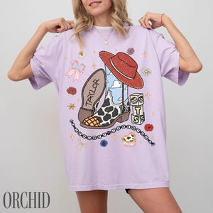 May include: A light purple short-sleeve t-shirt featuring a graphic print of a cowboy boot with a cow print pattern, a red hat, building blocks with the letters T, S, and the number 13, a pink bow, and a beaded string with the text THE ERAS TOUR.