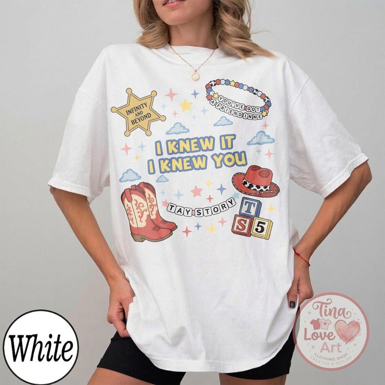 May include: A white short-sleeve t-shirt featuring a graphic print with the text I KNEW IT I KNEW YOU, TAY STORY, and INFINITY AND BEYOND. The design includes illustrations of a cowboy boot, a cowboy hat, alphabet blocks, and a beaded bracelet.