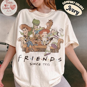 Peut inclure: Un t-shirt ivoire avec une impression graphique repr&eacute;sentant divers personnages anim&eacute;s assis sur un canap&eacute;. Sous les personnages, le texte indique "FRIENDS" avec des points color&eacute;s entre les lettres, suivi du texte "SINCE 1995" en noir.