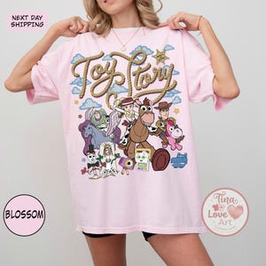 Op de afbeelding: Een lichtroze T-shirt met een grafische print van Toy Story-personages, waaronder Woody, Jessie, Buzz Lightyear, Bullseye, Forky en anderen. De tekst Toy Story staat in een touwachtig lettertype boven de personages tegen een blauwe lucht.