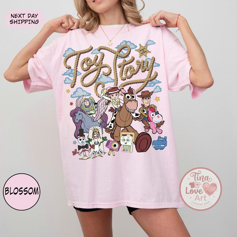 Op de afbeelding: Een lichtroze T-shirt met een grafische print van Toy Story-personages, waaronder Woody, Jessie, Buzz Lightyear, Bullseye, Forky en anderen. De tekst Toy Story staat in een touwachtig lettertype boven de personages tegen een blauwe lucht.
