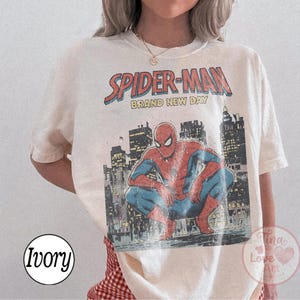 May include: Ivory-colored t-shirt featuring a vintage Spider-Man graphic. The design includes the words "SPIDER-MAN BRAND NEW DAY" above an illustration of Spider-Man crouching in front of a cityscape. The shirt has a classic crew neck.