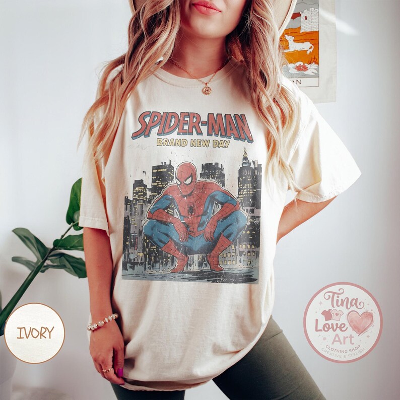 May include: Ivory-colored t-shirt featuring a vintage-style Spider-Man graphic. The design includes the words "SPIDER-MAN BRAND NEW DAY" above an illustration of Spider-Man crouching in front of a cityscape. The shirt has short sleeves and a relaxed fit.