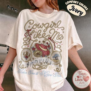 Pu&ograve; includere: Una t-shirt a maniche corte color avorio con una stampa grafica con il testo Cowgirl Like Me e Takes One to Know One. Il design include un cappello da cowboy rosso, un ferro di cavallo, un albero, note musicali e motivi a corda.