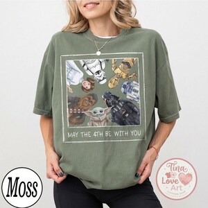 May include: Olive green t-shirt featuring a framed graphic of Star Wars characters including Princess Leia, Stormtrooper, C-3PO, R2-D2, Chewbacca, Darth Vader, and Grogu. The text "MAY THE 4TH BE WITH YOU" is below the graphic.