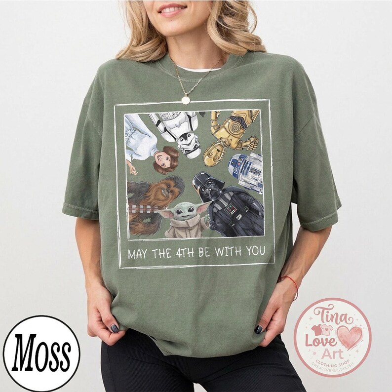 May include: Olive green t-shirt featuring a framed graphic of Star Wars characters including Princess Leia, Stormtrooper, C-3PO, R2-D2, Chewbacca, Darth Vader, and Grogu. The text "MAY THE 4TH BE WITH YOU" is below the graphic.
