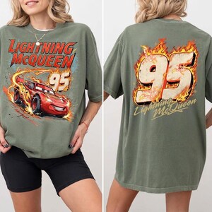 May include: An olive green t-shirt featuring a graphic of the red race car Lightning McQueen with the text Lightning McQueen and the number 95 on the front. The back displays a large number 95 surrounded by flames with the text Lightning McQueen.