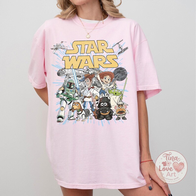 May include: Light pink t-shirt featuring a graphic of Star Wars characters in a cartoon style. The Star Wars logo is in yellow. Characters include Woody, Buzz Lightyear, and Darth Vader. The shirt has short sleeves.