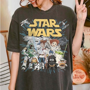 May include: Dark gray t-shirt featuring a Star Wars graphic with characters from the movie and Toy Story. The design includes the Star Wars logo in yellow, and images of Darth Vader, Woody, and other characters.