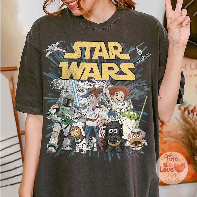 May include: Dark gray t-shirt featuring a Star Wars graphic with characters from the movie and Toy Story. The design includes the Star Wars logo in yellow, and images of Darth Vader, Woody, and other characters.